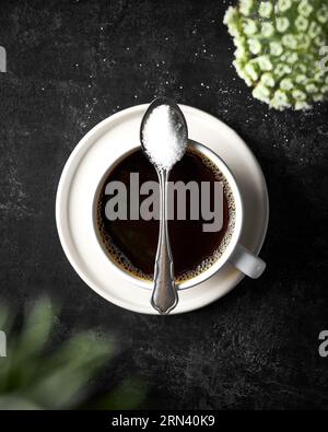 Top view picture of a black coffee in a white cup on a dark black textured background. Spoon of sugar on top of the cup. Blurry flowers in the foregro Stock Photo