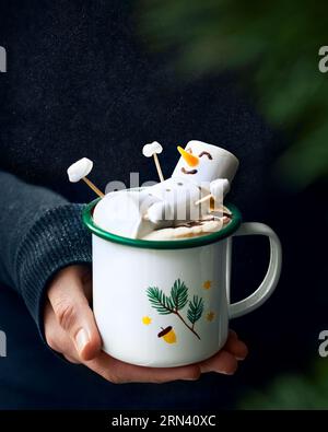 https://l450v.alamy.com/450v/2rn40xc/edible-marshmallow-snowman-laying-on-top-of-the-hot-cocoa-in-a-winter-themed-mug-a-womans-hand-holding-it-black-dark-background-2rn40xc.jpg