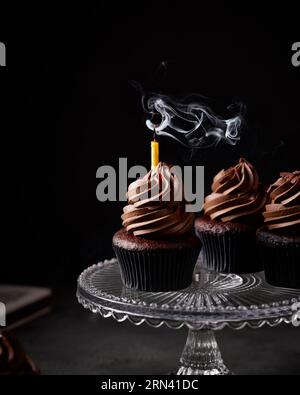 Three chocolate cupcakes on a glass cake stand. There is candle in one cupcake that has been blown out and there is still candle smoke visible. Stock Photo