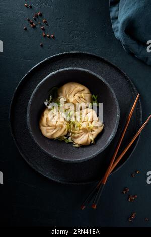 Ready to eat dim sum in the bowl garnished with herbs. Black ceramic textured bowl and plate on the black stone textured background. Moody light. Stock Photo