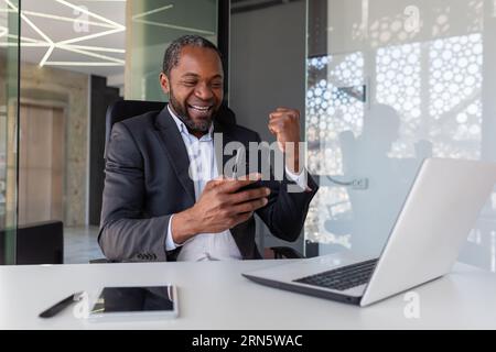 Mature successful african american businessman received online notification of winning message at workplace inside office, man holding phone, using app on smartphone, happy winner with achievement. Stock Photo