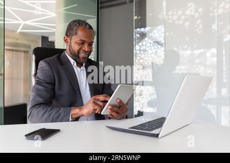 Mature adult african american boss investor at workplace using tablet computer, man happy with financial results and achievement smiling, using app inside office at table. Stock Photo