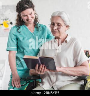 Nurse standing disabled senior woman reading book Stock Photo