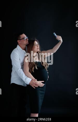 A young couple of parents-to-be taking selfies against at a black background Stock Photo