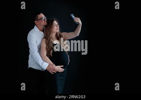 A young couple of parents-to-be taking selfies against at a black background Stock Photo
