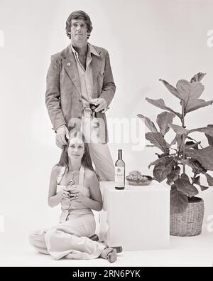 1970s FASHIONABLE COUPLE MAN STANDING WOMAN SITTING CUBE TABLE WITH WINE BOTTLE BOTH HOLDING GLASSES OF WINE LOOKING AT CAMERA - f13393 HAR001 HARS EYE CONTACT CUBE BEVERAGE STYLES JUMPSUIT FLUID HAIRSTYLE GRAPES HOUSE PLANT LIQUOR STYLISH SPIRITS BOOZE FASHIONS MID-ADULT MID-ADULT MAN MID-ADULT WOMAN SUEDE BEVERAGES BLACK AND WHITE CAUCASIAN ETHNICITY HAR001 OLD FASHIONED Stock Photo