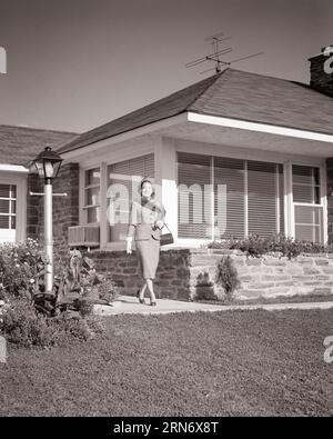 1950s WOMAN WALKING ON SIDEWALK IN FRONT OF MID-CENTURY MODERN HOUSE WEARING HAT WITH FEATHER A SUIT WHITE GLOVES MINK FUR PIECE - p1644 HAR001 HARS COPY SPACE FULL-LENGTH LADIES PERSONS RESIDENTIAL BUILDINGS MINK B&W BUSINESSWOMAN EYE CONTACT STRUCTURE STYLES EXTERIOR HOMES AIR CONDITIONER RESIDENCE STYLISH BUSINESSWOMEN IN FRONT OF FASHIONS MID-ADULT MID-ADULT WOMAN TV ANTENNA BLACK AND WHITE CAUCASIAN ETHNICITY HAR001 MID-CENTURY OLD FASHIONED Stock Photo