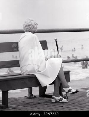 1970s 1980s ELDERLY WOMAN SITTING ON BOARDWALK BENCH WEARING WHITE SANDALS HOLDING CANE LOOKING OUT AT OCEAN - s21822 HAR001 HARS ELDER FEMALES CANE UNITED STATES COPY SPACE FULL-LENGTH LADIES PERSONS INSPIRATION UNITED STATES OF AMERICA SENIOR ADULT BOARDWALK B&W NORTH AMERICA SUMMERTIME SENIOR WOMAN NORTH AMERICAN RESORT SHORE DREAMS OLD AGE OLDSTERS OLDSTER LEISURE AGING RECREATION REAR VIEW BEACHES NJ ELDERS FROM BEHIND ESCAPE SANDY STYLISH DOWN THE SHORE NEW JERSEY OCEAN CITY BACK VIEW ELDERLY WOMAN RELAXATION SANDALS SEASON BLACK AND WHITE CAUCASIAN ETHNICITY COASTAL HAR001 OLD FASHIONED Stock Photo