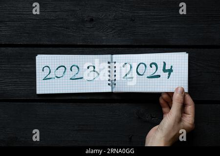 Notepad with the text 2023 and 2024 drawn with paint and a woman's hand turns over a page of a notebook on a black wooden board, the beginning of a ne Stock Photo