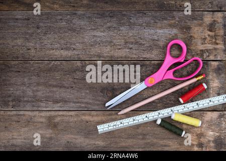 A flat, top view of a Sewing background with scissors, pencil, cotton reel threads and ruler in a rustic wooden setting with copy space to the left Stock Photo