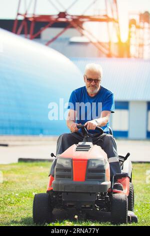 Worker mows territory of industrial facility. Man rides self-propelled lawn mower on lawn on summer sunny day. Elderly pensioner moonlights as gardener. Stock Photo