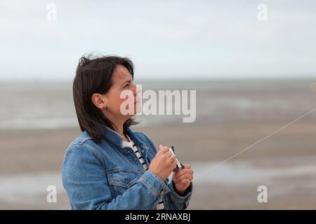 A middle-aged woman dressed in a denim jacket flies a kite on the beach at low tide. Bad weather during summer vacation Stock Photo