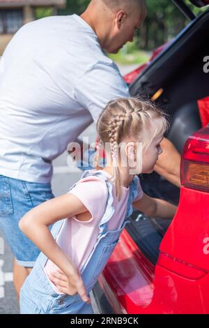 A girl with braids is putting bags in trunk of a red car next to her dad Stock Photo