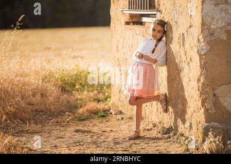 little girl in pink skirt leaning against rustic wall at golden hour Stock Photo