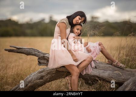 mother and daughter sitting on tree branch in open field hugging Stock Photo