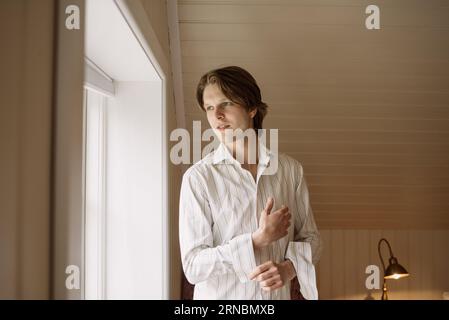 Happy young man standing near window and buttoning shirt sleeves Stock Photo