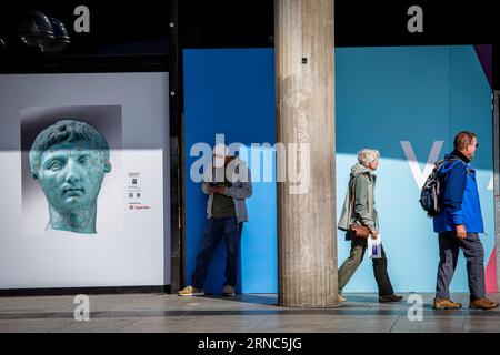 a man stands in front of the Romano-Germanic Museum next to a poster with a photo of the portrait head of Augustus, passerbys, Cologne, Germany. ein M Stock Photo