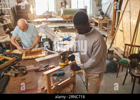 Portrait of young black man building wooden furniture while working in carpentry workshop and using electric tools, copy space Stock Photo