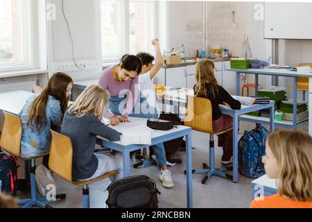 Female teacher assisting schoolgirls while sitting on desk in classroom Stock Photo