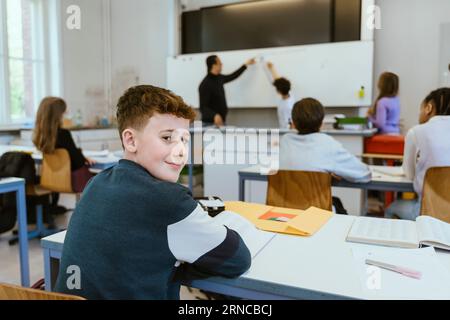 Portrait of smiling boy looking over shoulder while sitting on desk in classroom Stock Photo