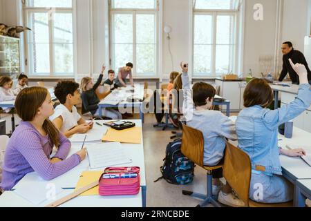 Male and female students raising hands while sitting on chairs in classroom Stock Photo