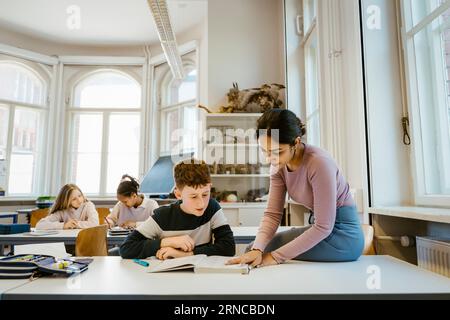 Female teacher assisting schoolboy reading book while sitting at desk in classroom Stock Photo