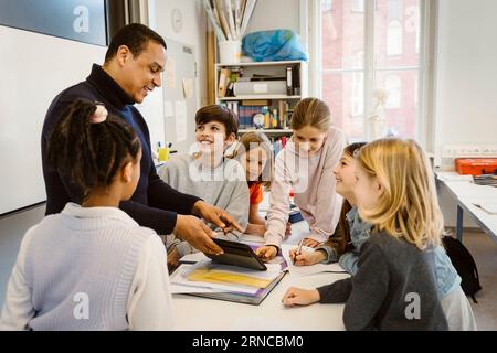 Male teacher teaching students through tablet PC at desk in classroom Stock Photo