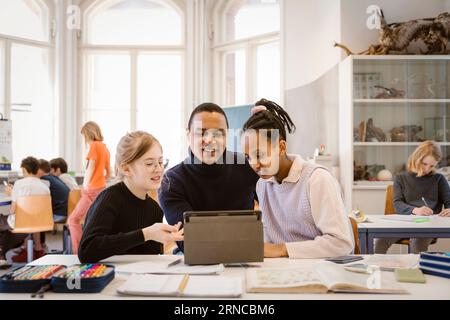 Smiling male teacher teaching female students through tablet PC in classroom Stock Photo