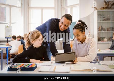 Smiling male teacher doing E-learning with female students in classroom Stock Photo