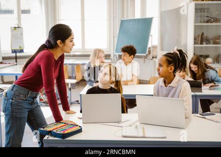 Female teacher talking to schoolgirls sitting with laptops on desk in classroom Stock Photo