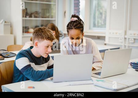 Boy sharing laptop with female friend sitting at desk in classroom Stock Photo