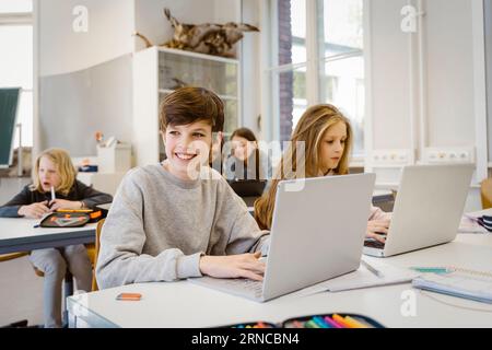 Happy boy looking away while sitting with laptop by female friend at desk in classroom Stock Photo