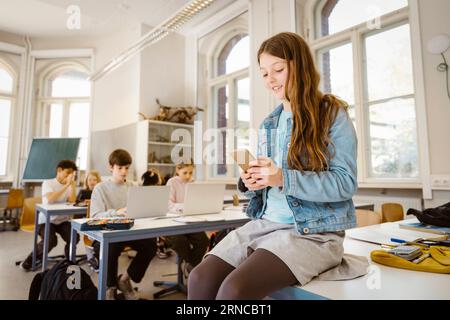 Smiling female student using smart phone while sitting at desk in classroom Stock Photo