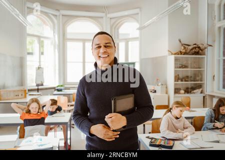 Portrait of smiling male teacher holding tablet PC while standing in classroom Stock Photo