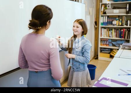 Smiling female student talking to teacher standing near whiteboard in classroom Stock Photo