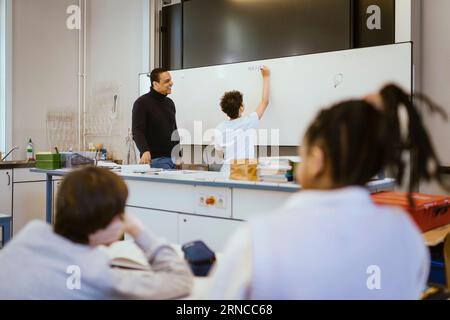 Smiling teacher standing with schoolboy writing on whiteboard in classroom Stock Photo