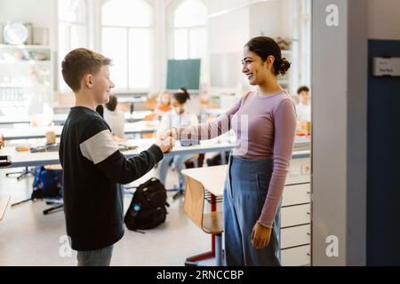 Smiling teacher shaking hand with schoolboy standing in classroom Stock Photo