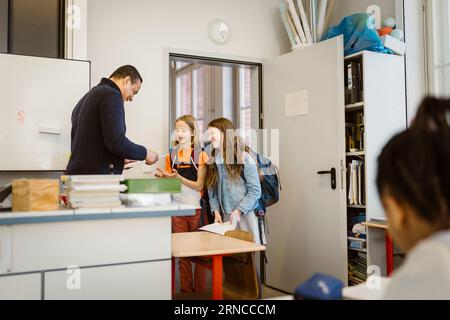 Mature male teacher giving documents to students standing in classroom Stock Photo
