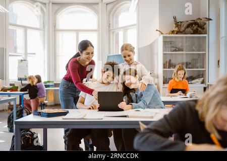 Smiling male and female students sharing tablet PC with teacher in classroom Stock Photo