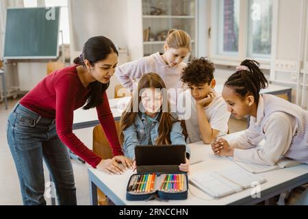 Male and female students doing E-learning with teacher leaning at desk in classroom Stock Photo