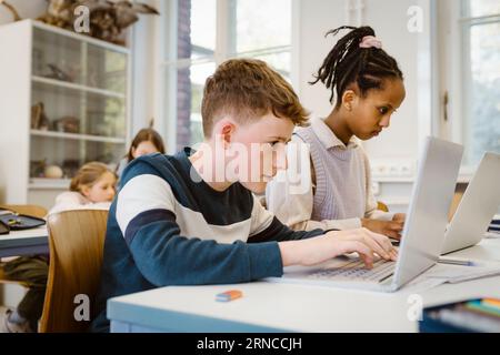 Focused schoolboy using laptop by female friend at desk in classroom Stock Photo