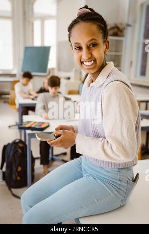 Portrait of happy schoolgirl holding smart phone while sitting at desk in classroom Stock Photo