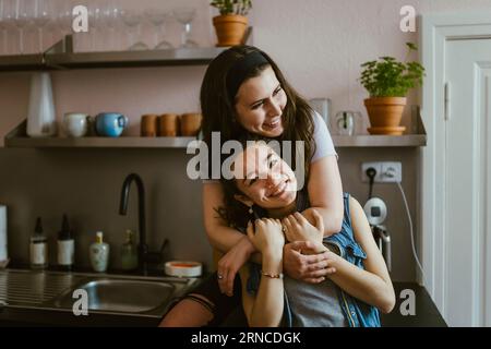 Portrait of happy woman embracing female friend from behind while sitting in kitchen at home Stock Photo
