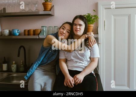 Young woman hugging sister with down syndrome in kitchen at home Stock Photo