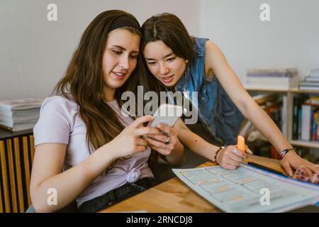 Young female friends sharing smart phone while studying together at home Stock Photo