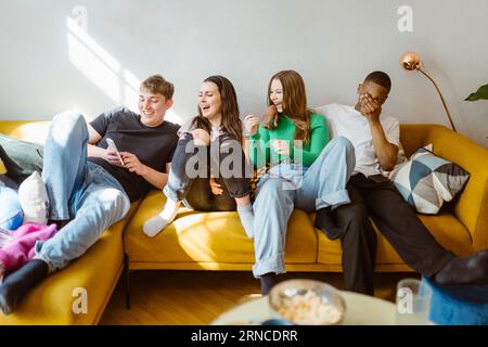 Happy young multiracial friends using smart phone while sitting together on sofa at home Stock Photo