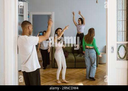 Young man photographing female friends dancing in living room at home Stock Photo