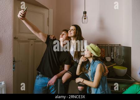 Young man taking selfie with smiling female friends in kitchen at home Stock Photo
