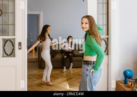 Portrait of smiling young woman standing at doorway near friends dancing in living room Stock Photo