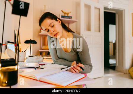 Young woman reading book while doing homework at table Stock Photo